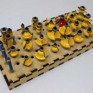 May include: A rectangular wooden storage rack with interlocking edges, featuring numerous holes holding a collection of various router bits. The bits have yellow, blue, or red bodies with silver metal cutting edges and ball-bearing guides.