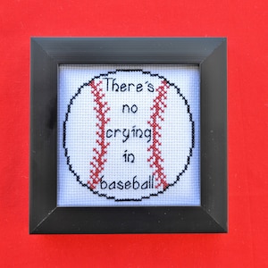 May include: A black framed cross-stitch with a white background and a red baseball with the text "There's no crying in baseball" in black.