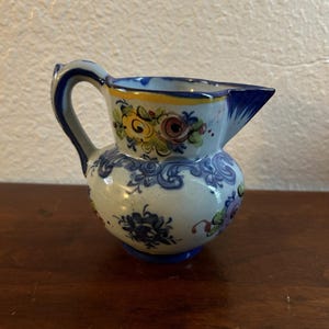 May include: A small, hand-painted ceramic pitcher with a rounded body and a spout. The pitcher features a white base with blue floral patterns and colorful flowers in yellow, pink, and green. The handle and rim are accented with blue and yellow.