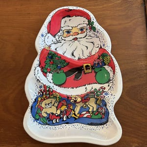 May include: A white ceramic serving tray shaped like Santa Claus. The design features a central illustration of Santa with a red hat, white beard, and green mittens, holding a decorated Christmas tree and gifts. The base shows toys including a teddy bear, a rocking horse, and a train.
