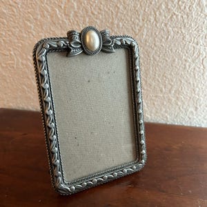 May include: A rectangular silver-toned picture frame with a bow and oval embellishment at the top. The frame has a decorative border and a neutral-colored backing. The frame is displayed on a wooden surface.
