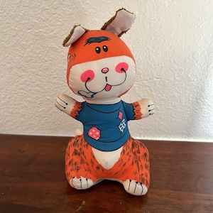 May include: A vintage plush dog toy with an orange head and body, brown ears, and a blue shirt. The dog has a white face with pink cheeks and a smiling mouth. The toy is sitting on a wooden surface.