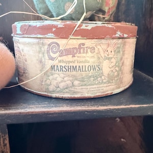 May include: A vintage Campfire Whipped Vanilla Marshmallows tin with a distressed, rustic appearance. The tin features a red and teal lid, with a bunny illustration and text. The tin is cylindrical and sits on a dark wooden shelf.