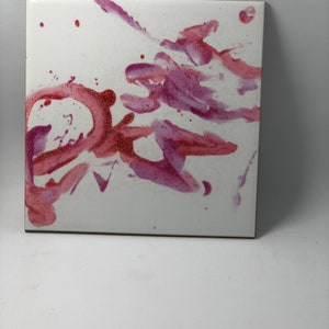 Puede incluir: Un azulejo de cer&aacute;mica blanco con salpicaduras abstractas de pintura rosa y roja. La pintura tiene una textura brillante.