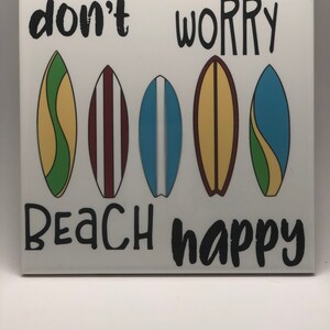 Puede incluir: Azulejo de cer&aacute;mica blanco con el texto "don't worry beach happy" en negro. Cinco ilustraciones de tablas de surf coloridas est&aacute;n dispuestas en una fila sobre el texto.