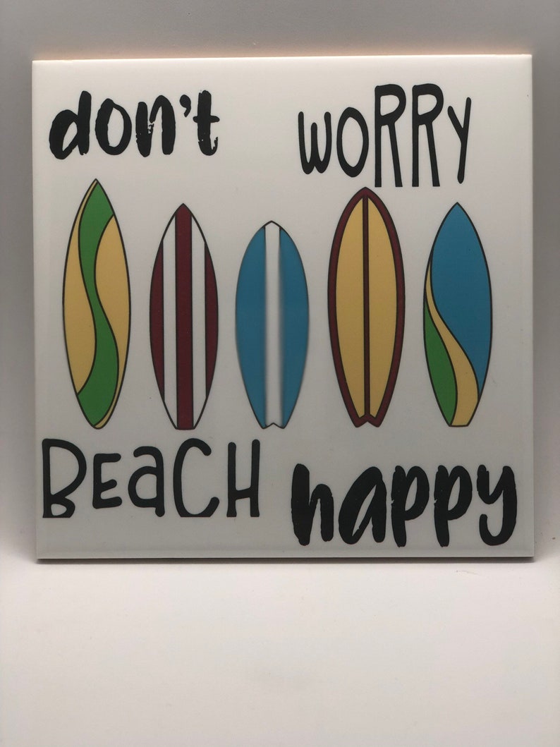 Puede incluir: Azulejo de cer&aacute;mica blanco con el texto "don't worry beach happy" en negro. Cinco ilustraciones de tablas de surf coloridas est&aacute;n dispuestas en una fila sobre el texto.
