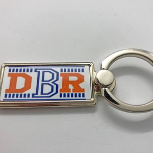 May include: A silver keychain with a rectangular tag featuring the letters "DBR" in orange and blue on a white background. The tag has a blue and white striped border.
