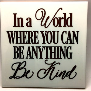 Puede incluir: Azulejo de cer&aacute;mica blanco con el texto "In a world where you can be anything Be Kind" en letras marrones.