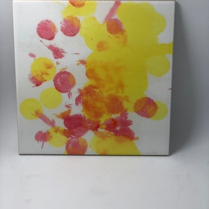 Puede incluir: Un azulejo de cer&aacute;mica blanco con salpicaduras abstractas de pintura amarilla y roja. La pintura est&aacute; en un patr&oacute;n circular, creando un dise&ntilde;o din&aacute;mico y colorido.