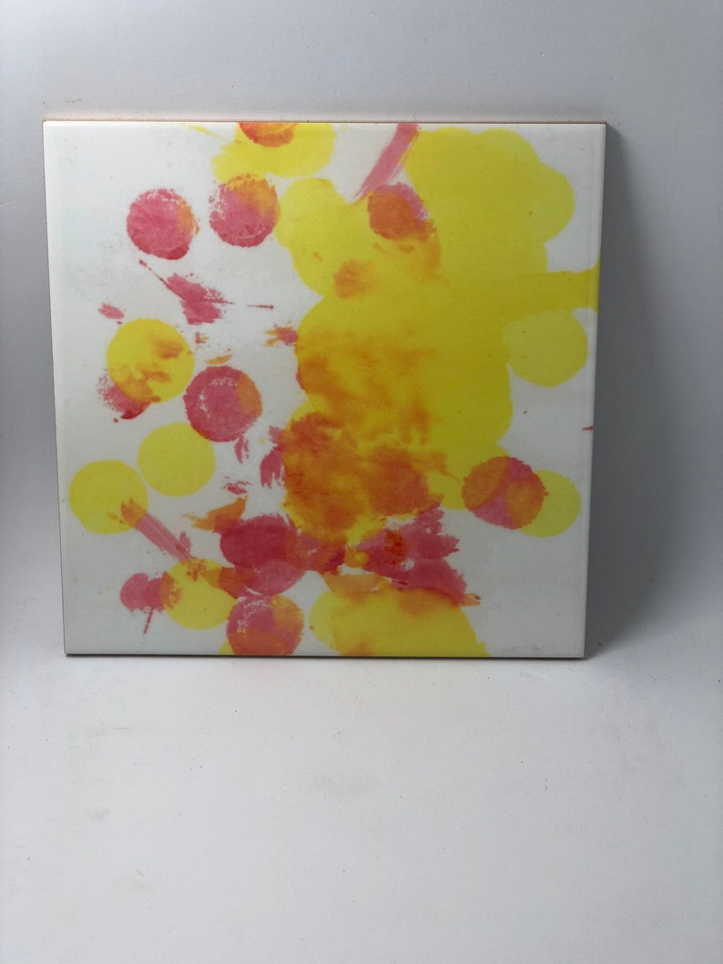 Puede incluir: Un azulejo de cer&aacute;mica blanco con salpicaduras abstractas de pintura amarilla y roja. La pintura est&aacute; en un patr&oacute;n circular, creando un dise&ntilde;o din&aacute;mico y colorido.