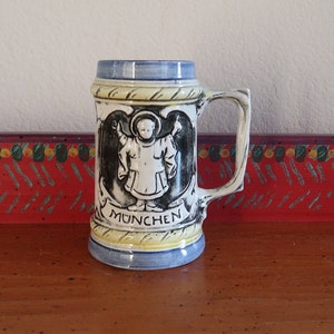 May include: A ceramic beer stein with a blue and yellow design. The stein features a black and white image of a cherub with wings and the word "MUNCHEN" on the front.