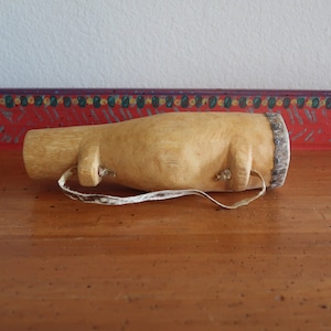 May include: A wooden, cylindrical musical instrument with a leather strap. The instrument is light brown and has a natural wood finish.