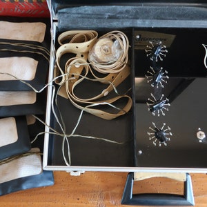 May include: A black case with a silver handle and a black interior. The case contains a set of beige pads, a beige strap, and a black control panel with knobs and switches. The control panel has the text "ON BATTERY OFF" and a logo with three figures.