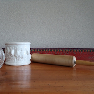 Könnte beinhalten: Ein weißer Keramikbehälter mit einem erhabenen Design, eine klare Glasvase und ein Nudelholz aus Holz auf einer Holzoberfläche.