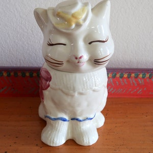 Pode incluir: Jarro de biscoitos de cerâmica branca em forma de gato com uma flor amarela na cabeça, uma flor rosa no lado e uma borda azul e branca ao redor do pescoço. O gato tem olhos castanhos e um sorriso fechado.