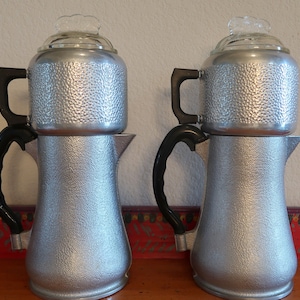 May include: Two silver percolator coffee pots with black handles and glass lids. The pots have a textured, hammered finish.