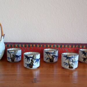 May include: A set of seven ceramic teacups and a teapot with a bamboo design. The cups and teapot are white with blue and green accents. The teapot has a bamboo handle.