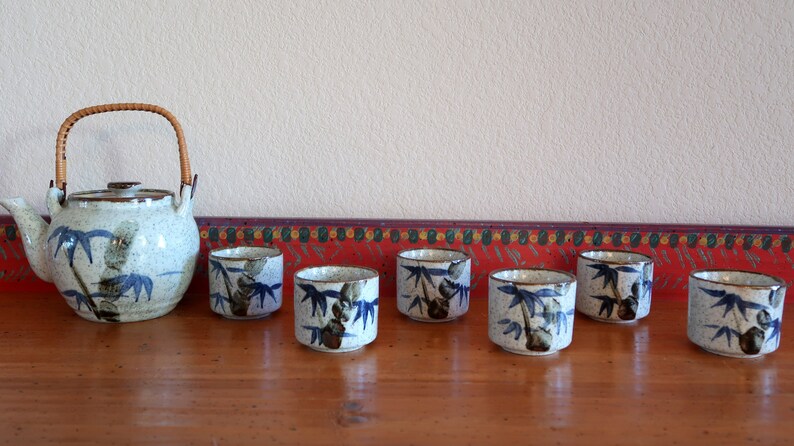 May include: A set of seven ceramic teacups and a teapot with a bamboo design. The cups and teapot are white with blue and green accents. The teapot has a bamboo handle.