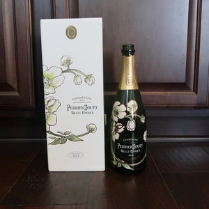 Könnte beinhalten: Eine Flasche Perrier-Jouët Belle Epoque Champagner mit goldenem Etikett und floralem Design. Die Flasche steht auf einer Holzoberfläche neben einer Schachtel mit dem gleichen Design.