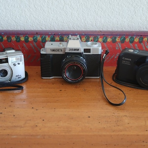 May include: Three different film cameras on a wooden surface. The camera on the left is silver and says "PENTAX" on the front. The camera in the middle is black and silver and says "YAMASHETA 35MM" on the front. The camera on the right is black and says "Canon Sure Shot ZOOM 110" on the front.
