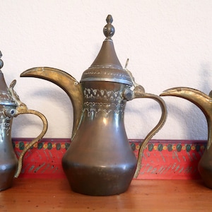 May include: Three antique brass coffee pots with ornate designs and lids. The pots are arranged in a row on a wooden surface.