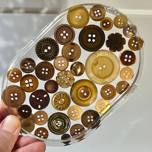 Può includere: Sottobicchiere ovale in resina con una varietà di bottoni marroni e beige. I bottoni variano per dimensioni, forma e materiale, tra cui plastica e metallo, creando una superficie unica e strutturata. Il sottobicchiere è tenuto da una mano.
