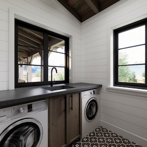 May include: A modern laundry room with white shiplap walls and a dark countertop. Features include a black sink and faucet, two front-load washing machines, and a wooden cabinet. Black-framed windows offer natural light and outdoor views.
