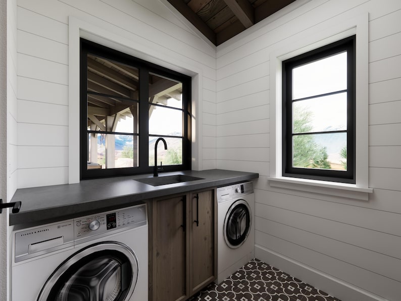 May include: A modern laundry room with white shiplap walls and a dark countertop. Features include a black sink and faucet, two front-load washing machines, and a wooden cabinet. Black-framed windows offer natural light and outdoor views.