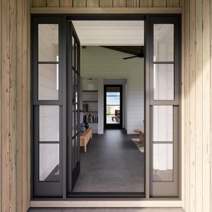 May include: An open doorway with black-framed glass doors and windows, leading into a well-lit interior. The exterior features light-colored wooden siding. The interior reveals a living space with a bench, bookshelves, and a view of the outdoors.