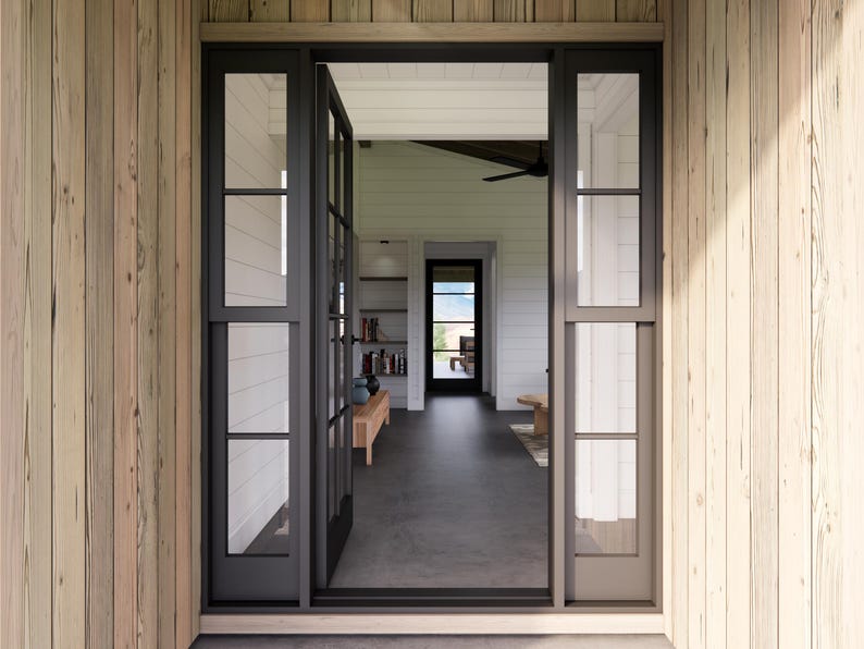 May include: An open doorway with black-framed glass doors and windows, leading into a well-lit interior. The exterior features light-colored wooden siding. The interior reveals a living space with a bench, bookshelves, and a view of the outdoors.