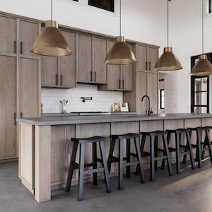 May include: A modern kitchen with light wood cabinetry, a gray countertop, and black bar stools. Three pendant lights with gold-colored shades hang above the island. The kitchen features a white tile backsplash and a black faucet.