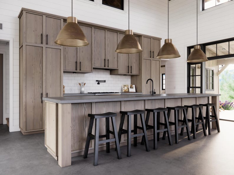 May include: A modern kitchen with light wood cabinetry, a gray countertop, and black bar stools. Three pendant lights with gold-colored shades hang above the island. The kitchen features a white tile backsplash and a black faucet.