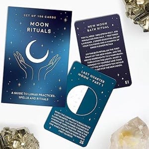 May include: A set of 100 Moon Rituals cards featuring a dark blue box with a white crescent moon and hands illustration. Two individual cards are displayed: one titled New Moon Bath Ritual and another titled Last Quarter Moon - Part 1.