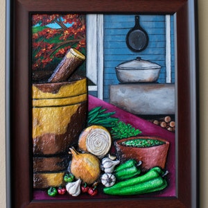 Puede incluir: Una pintura enmarcada que representa una naturaleza muerta de ingredientes de cocina. Un mortero y un pilón, cebollas, ajos, pimientos verdes y un bol de salsa verde están dispuestos sobre un mantel rojo. El fondo muestra una cocina azul y blanca con una olla y una sartén colgadas en la pared.