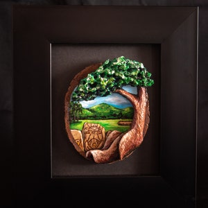 May include: A framed, three-dimensional artwork depicting a landscape scene. The artwork features a tree with green foliage, a grassy field, rolling green hills, and a carved stone. The frame is black.