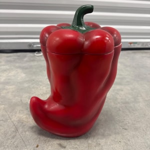 May include: A glossy red ceramic cookie jar shaped like a bell pepper with a dark green stem lid. The container features a curved, bulbous body with deep indentations and a smooth, reflective finish, standing upright as a functional kitchen storage piece.