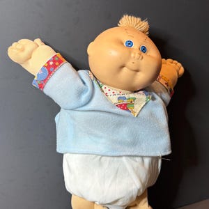 May include: A soft-bodied doll with a round vinyl head, blue eyes, and a small tuft of blonde yarn hair. The doll wears a light blue long-sleeved shirt with a colorful patterned collar and sleeve cuffs, paired with white fabric diaper-style bottoms.