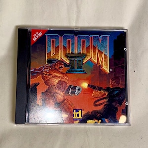 May include: A clear plastic jewel case for the video game Doom II for the Macintosh. The cover art features a large horned demon facing a soldier firing a weapon, with the title Doom II in large gold letters and the id Software logo below.