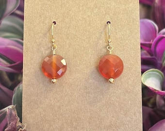 Może przedstawiać: Para pomarańczowych kolczyków z kamieni szlachetnych ze złotymi haczykami wisi na brązowej etykiecie. Na etykiecie widnieje napis "VERMONT CREATION HANDCRAFTED BY BETH KURASH". Kolczyki są umieszczone na tle fioletowych i zielonych roślin.