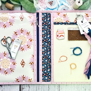 May include: A pink and white fabric project organiser with a zipper closure. The organiser has a pocket for scissors, needles, and thread. It also has a pocket for a small milk carton-shaped pincushion with a rainbow and heart design. The organiser has a blue and white floral fabric trim.