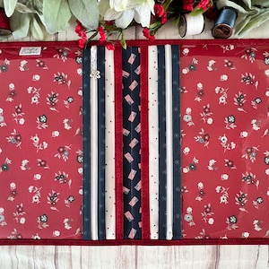 Puede incluir: Una bolsa de tela roja con cremallera y ventanas de plástico transparente, decorada con cintas y tiras de tela blancas y azules. La tela tiene un pequeño estampado floral blanco.