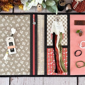 Peut inclure: Un organisateur enroulable en tissu noir avec plusieurs poches et compartiments. L'organisateur a un imprimé floral, un imprimé journal, un imprimé vichy et un tissu rose uni. L'organisateur comprend des aiguilles, du fil et d'autres fournitures de couture. Le texte "Wool Knits" est visible sur le tissu rose.