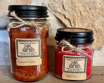 May include: Two glass jar candles with black lids and twine bows. One candle is filled with a brown wax and labeled "Toasted Hazelnut." The other is filled with red wax and labeled "Apple Spice." Both labels read "Cabin Fever Primitives."