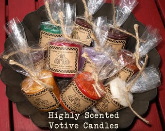 May include: A collection of multi-colored votive candles individually wrapped in clear plastic and tied with twine. Each candle features a tan paper label with the text Cabin Fever Primitives and a line drawing of a cabin. The candles are arranged in a dark metal bowl.
