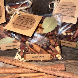 May include: A clear plastic bag filled with dried potpourri ingredients including cinnamon sticks, bay leaves, and spices. A brown paper tag labeled Apple Spice is attached with twine, featuring instructions to add a heaping Tbsp to boiling water.