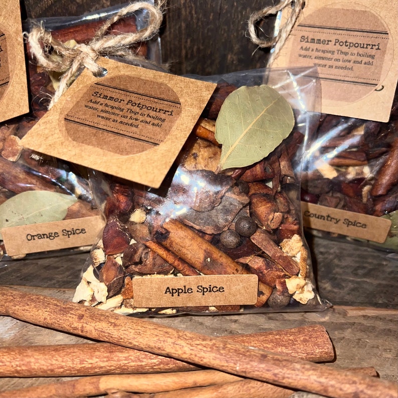 May include: A clear plastic bag filled with dried potpourri ingredients including cinnamon sticks, bay leaves, and spices. A brown paper tag labeled Apple Spice is attached with twine, featuring instructions to add a heaping Tbsp to boiling water.