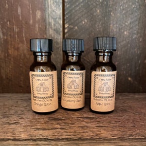 May include: Three small brown glass bottles with black caps. Each bottle has a label with the text "Cabin Fever" and "Primitives". The labels read "Orange Spice", "Cranberry Jam", and "Apple Spice".