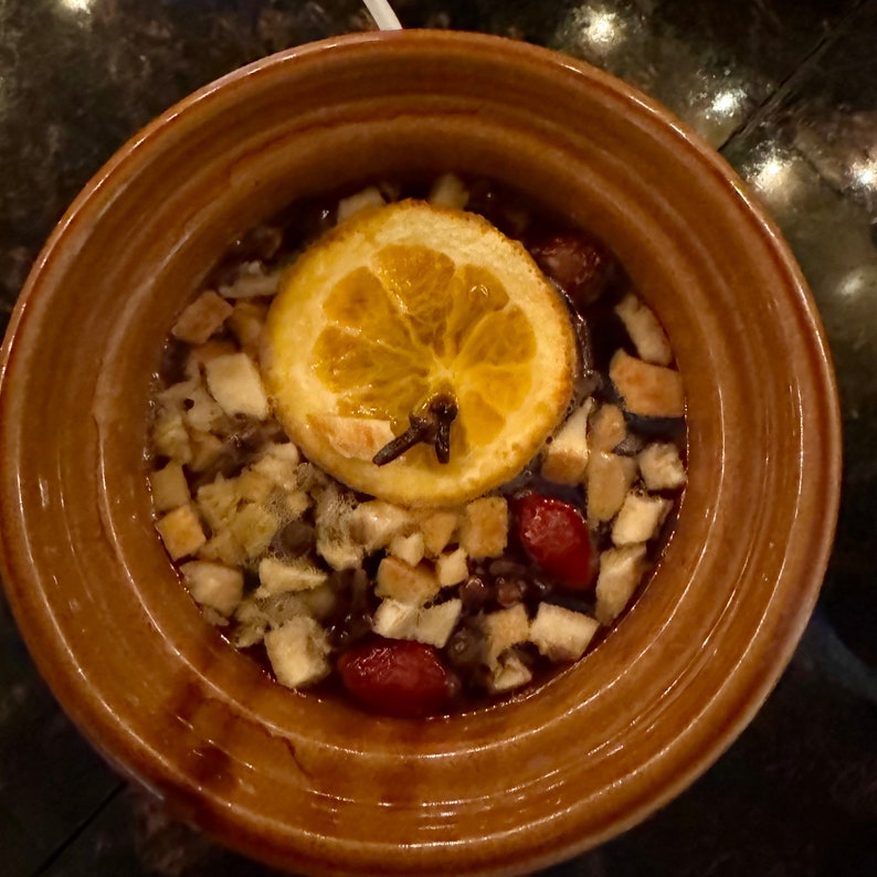 May include: A round, brown ceramic bowl filled with a dark liquid, topped with a single dehydrated orange slice, a whole clove, small pieces of dried bread or fruit, and several whole red berries, viewed from directly above.