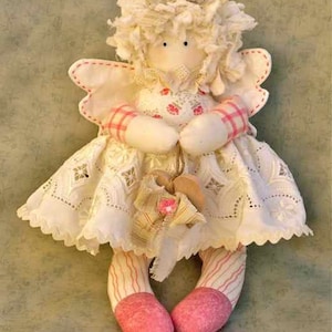 Può includere: Una bambola di angelo di stoffa bianca con gambe a righe rosa e bianche e scarpe rosa. La bambola indossa un vestito bianco con bordino di pizzo e un corpetto a quadri rosa e bianco. La bambola ha un'aureola di stoffa bianca e un fiocco di stoffa bianca.