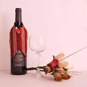 May include: A red and gold striped wine bottle with a bow tie and vest. A clear wine glass and a bouquet of cream and red roses are on a white surface.